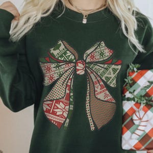 May include: A forest green crewneck sweatshirt featuring a large central graphic of a bow. The bow is composed of various patterned sections in red, white, and green, including snowflakes, reindeer, and geometric shapes. The text Forest green is visible.