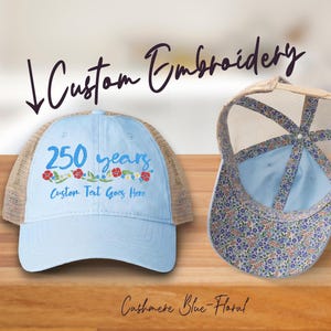 May include: Two light blue trucker hats with custom embroidery. One hat features "250 years" in blue with floral accents and "Custom Text Goes Here." The other hat has a floral pattern on the underside of the brim. The text "Custom Embroidery" is above the hats.