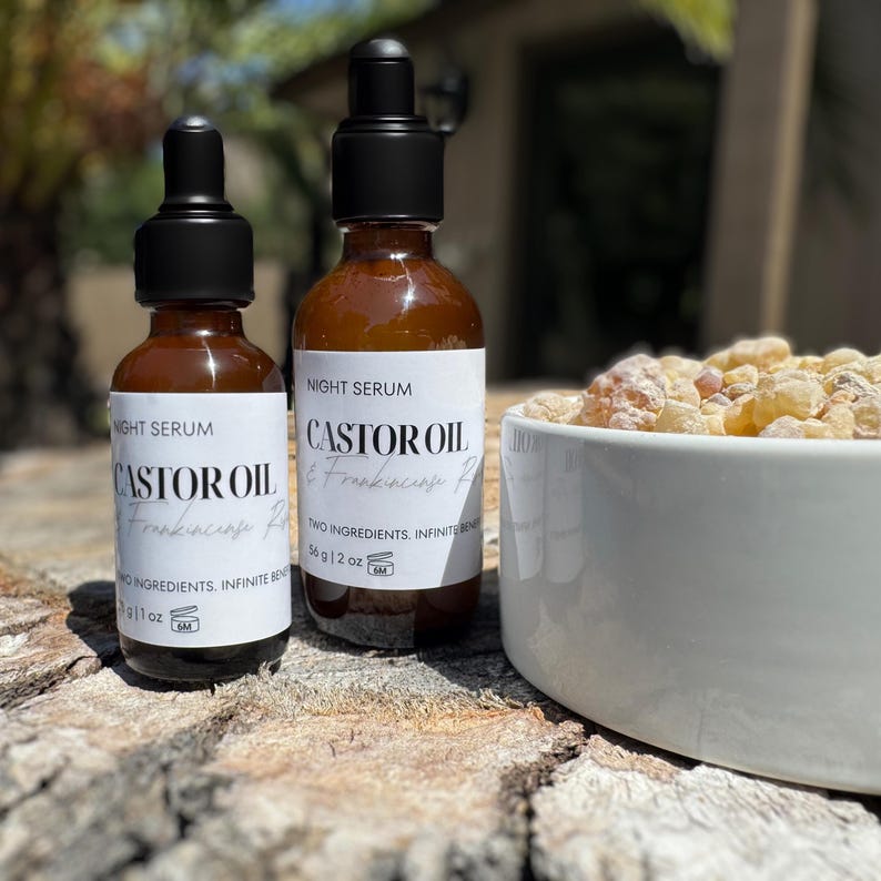 May include: Two amber glass bottles of night serum with black droppers, labeled "Castor Oil & Frankincense." The bottles are on a wooden surface next to a white bowl filled with light-colored resin. The labels also state "Two Ingredients. Infinite Benefits."