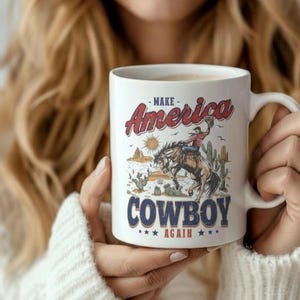 May include: White ceramic mug featuring a cowboy riding a bucking bronco, with desert scenery and the text "MAKE America COWBOY AGAIN." The mug has a curved handle and is held by a person wearing a white sweater.