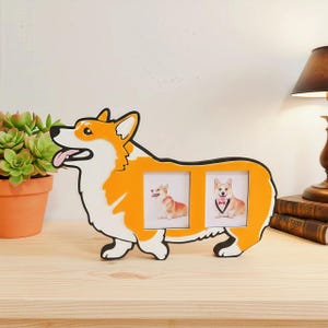 May include: A corgi-shaped photo frame in orange, white, and black. The frame holds two photos of corgis. The frame is on a wooden surface, with a potted plant and a lamp in the background. The frame is approximately 12 inches long.