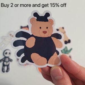 Könnte beinhalten: Ein Aufkleber mit einem Cartoon-Bären, der als Spinne verkleidet ist, mit einem schwarzen Körper und einem beigen Gesicht und Gliedmaßen. Der Aufkleber wird in einer Hand gehalten, mit anderen Halloween-Themenaufklebern im Hintergrund. Der Text oben lautet "Buy 2 or more and get 15% off."