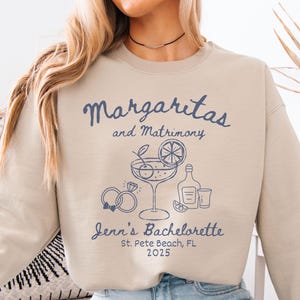 May include: Tan colored sweatshirt with a blue graphic design that says "Margaritas and Matrimony" with a margarita glass, lime, cherries, rings, and bottles. The text "Jenn's Bachelorette St. Pete Beach, FL 2025" is also included in the design.