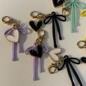 May include: A collection of heart-shaped charms with gold-colored clasps. The charms feature black, white, and lavender hearts, some with beaded edges, and attached bows in various colors. The charms are designed for use as keychains or accessories.