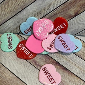 May include: A collection of heart-shaped candies in various colors, including pink, red, blue, and purple. Each candy has the word "SWEET" printed on it in a contrasting color. The candies are scattered on a wooden surface.