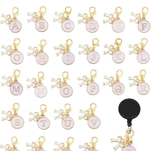 May include: A collection of gold-toned alphabet charms with white enamel backgrounds. Each charm features a letter from A to Z, attached to a lobster clasp and a decorative bow. A black retractable badge reel with the letter "I" is also included.