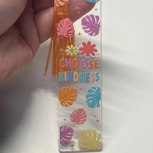 May include: Clear acrylic bookmark with colorful leaf and flower designs. The bookmark has the words "CHOOSE KINDNESS" in rainbow colors. It is adorned with an orange tassel and a gold string.