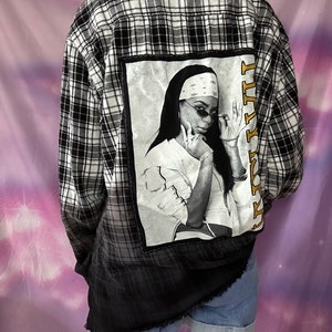 May include: Black and white plaid flannel shirt with a black and white photo of a woman wearing sunglasses and a bandana on the back. The photo has the text "Lil Kim" written vertically in gold letters.