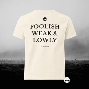 Könnte beinhalten: Cremefarbenes T-Shirt mit dem schwarzen Schriftzug „FOOLISH WEAK & LOWLY“, einem Totenkopf-Motiv und dem Bibelvers 1 Kor 1,27-29. Der Hintergrund zeigt eine verschwommene, monochrome Landschaft.
