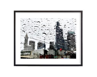 May include: A blurry view of a city skyline through a rain-streaked window. Raindrops are visible on the glass, and the buildings in the background are out of focus.
