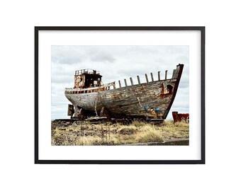 May include: A weathered wooden boat sits on land, partially submerged in the sand. The boat is rusty and has a faded blue paint job. The boat is surrounded by dry grass and a cloudy sky.