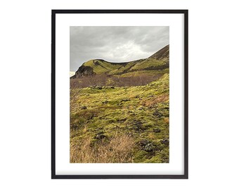 May include: A photograph of a green, mossy hillside with a rocky mountain in the background. The sky is overcast and the landscape is muted in color.