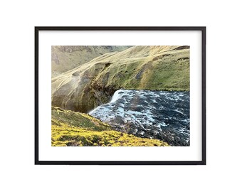 May include: A scenic landscape photograph of a waterfall cascading down a rocky cliff into a river. The water is a vibrant blue and white, and the surrounding hills are covered in lush green grass.
