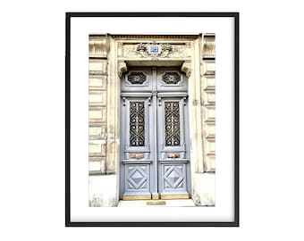 May include: A vintage-style, double-door entrance with a gray painted wood door and ornate ironwork. The door has a brass door knocker and a brass doormat. The building is made of light-colored stone with a decorative stone archway above the door. The number "112" is visible on the building.