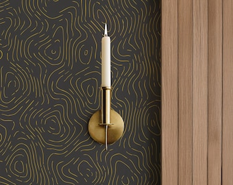 May include: A brass candle holder with a lit white candle. The candle holder is mounted on a dark gray wall with a gold line pattern. To the right is a wooden panel wall.