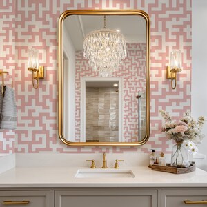 May include: A bathroom with a gold-framed mirror, a crystal chandelier, and gold fixtures. The walls are decorated with a pink and white geometric pattern. A white countertop holds a sink, soap dispensers, and a vase of flowers.