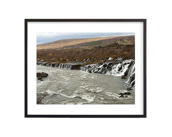 May include: A wide shot of a river flowing through a rocky landscape. The river is whitewater and there are several small waterfalls in the background. The landscape is covered in brown grass and trees.
