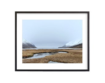 May include: A photograph of a landscape with a winding river flowing through a field of brown grass. The river leads to a body of water in the distance, framed by snow-capped mountains on either side. The sky is a pale blue with a hint of gray.