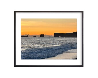 May include: A framed landscape print featuring a coastal scene at sunset. The image shows a beach with waves, rock formations, and a vibrant orange and yellow sky. The frame is black.