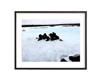 May include: A photograph of a blue lagoon with black rocks in the foreground. The water is a light blue color and the sky is overcast.