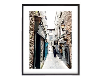 May include: A narrow, cobblestone alleyway in a European city. The alley is lined with old stone buildings, some with shops and restaurants. The buildings are painted in shades of gray and brown. A single person walks down the alleyway.
