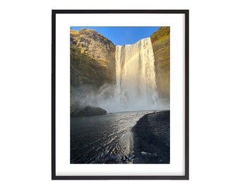 May include: A waterfall cascades down a rocky cliff face, creating a rainbow in the mist. The water flows into a dark, shallow pool at the base of the falls.