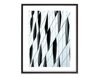 May include: Framed architectural print featuring a modern building facade. The image showcases a repeating pattern of angled, reflective panels in shades of white, gray, and black, creating a dynamic, abstract design. The frame is black.