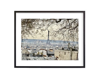 May include: Framed print of the Eiffel Tower in Paris, France, viewed from an elevated perspective. The black frame surrounds a cityscape with rooftops, buildings, and the iconic tower. Bare tree branches frame the top of the image.