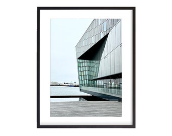 May include: A black and white photo of a modern building with a geometric facade. The building is situated on a waterfront with a wooden deck in the foreground.