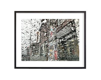 May include: A blurry image of a city street seen through a car window covered in raindrops. The raindrops are out of focus and the buildings in the background are blurred.
