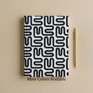 Modern abstract patterned hardcover journal mockup on a warm neutral background with a gold pen beside the journal and text reading “More Colors Available.” Charcoal and cream winding line pattern design with a clean modern aesthetic.