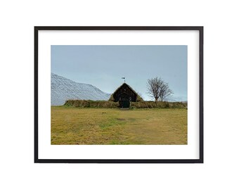 May include: A black wooden church with a grass roof stands in a field with a snow-capped mountain in the background.