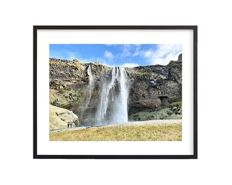 May include: A large waterfall cascades down a rocky cliff face, with a few people standing at the base. The sky is blue with white clouds.