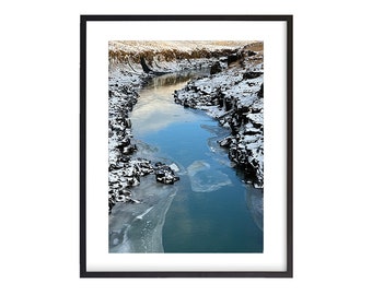以下が含まれることがあります： 凍った川が岩の多い渓谷を流れ、岩は雪で覆われ、水は青い空を映し出しています。