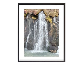 May include: A waterfall cascades down a rocky cliff face, with lush green foliage at the top. The water is clear and white, creating a dramatic contrast against the dark gray rock.