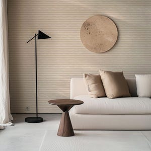 May include: A contemporary living room setting with a dark wood side table, featuring a conical base and a circular top. A black floor lamp with a triangular shade is positioned nearby. A beige sofa with cushions and a round wall art piece are also present.