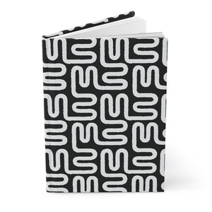Puede incluir: Un cuaderno de tapa dura con un patr&oacute;n geom&eacute;trico en blanco y negro. El patr&oacute;n consiste en formas blancas, estilizadas e interconectadas sobre un fondo negro. El cuaderno est&aacute; abierto, revelando p&aacute;ginas blancas en blanco.
