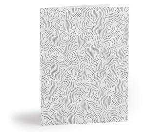 May include: A folded greeting card featuring a light gray background with a repeating pattern of thin, black, wavy, concentric lines that resemble a topographic map. The card is shown from a slight angle, revealing its folded edge and structure.