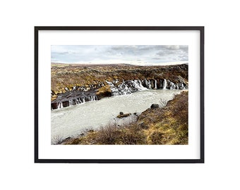 May include: A scenic landscape photograph of a waterfall cascading down a rocky cliff into a river. The water is a milky white color and the surrounding area is covered in brown and green vegetation.