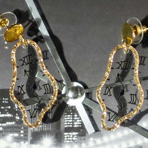 May include: A pair of gold-colored earrings with a clear, teardrop-shaped design. The edges are outlined with gold glitter. Roman numerals are printed on the clear surface. The earrings are set against a black and white cityscape background.