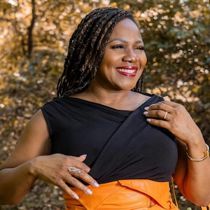 May include: A woman wearing a black top with an orange skirt, smiling. She is wearing a gold bracelet and ring. Her hair is styled in braids. The background is a blurred view of trees.