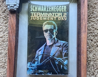 Può includere: Un nastro VHS incorniciato del film Terminator 2: Il giorno del giudizio con Arnold Schwarzenegger. Il nastro è in una cornice di legno con pezzi d'angolo decorativi.