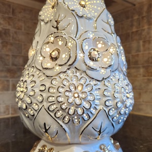 Puede incluir: Lámpara decorativa de cerámica blanca con diseño floral y detalles dorados. La lámpara tiene forma de árbol con intrincados patrones de flores y hojas. La base es cuadrada con adornos florales dorados. La lámpara está iluminada, con luz visible a través de los recortes florales.