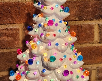May include: A white ceramic Christmas tree with colorful, round lights and a blue star topper. The tree is illuminated, showcasing a festive holiday decoration. The base is a matching white color.