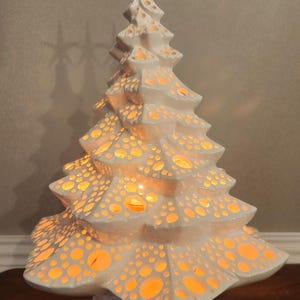 May include: A white ceramic Christmas tree with a star topper. The tree has numerous cut-out holes that emit a warm, orange glow when lit. The base is a decorative, white, bow-shaped design. The tree is approximately 12 inches tall.