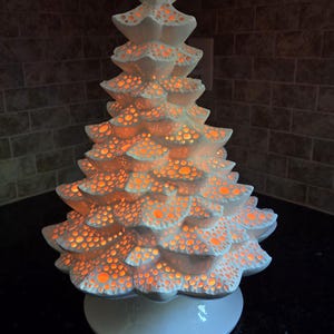 May include: A white ceramic Christmas tree with a textured, perforated design. The tree is illuminated from within, emitting a warm orange glow. The base is a matching white color. The text "Celebrate Ceramics" is at the bottom.