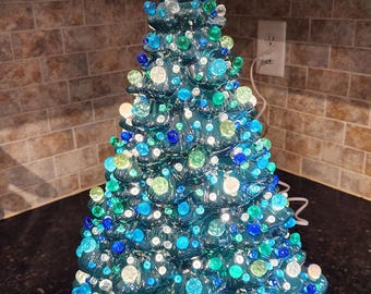 May include: A ceramic Christmas tree in shades of teal, blue, and green, adorned with decorative lights and a star topper. The tree is approximately 14 inches tall and sits on a dark surface.