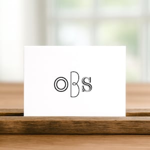 Puede incluir: Una tarjeta blanca con las letras "oBs" en una fuente decorativa, colocada sobre una superficie de madera. El fondo está borroso, lo que sugiere un entorno natural. La tarjeta es probablemente un artículo de papelería personalizado.