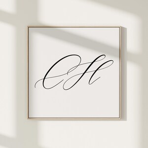 以下が含まれることがあります： 白い背景に黒い筆記体文字「C」と「H」が描かれた正方形の額入りプリント。アートワークは細い金色のフレームに収められており、ミニマリストな美学を作り出しています。プリントは白い壁に表示されています。