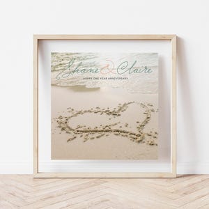 May include: Framed print featuring a beach scene with a heart drawn in the sand. The names "Shane & Claire" are written in elegant script above the text "Happy One Year Anniversary."
